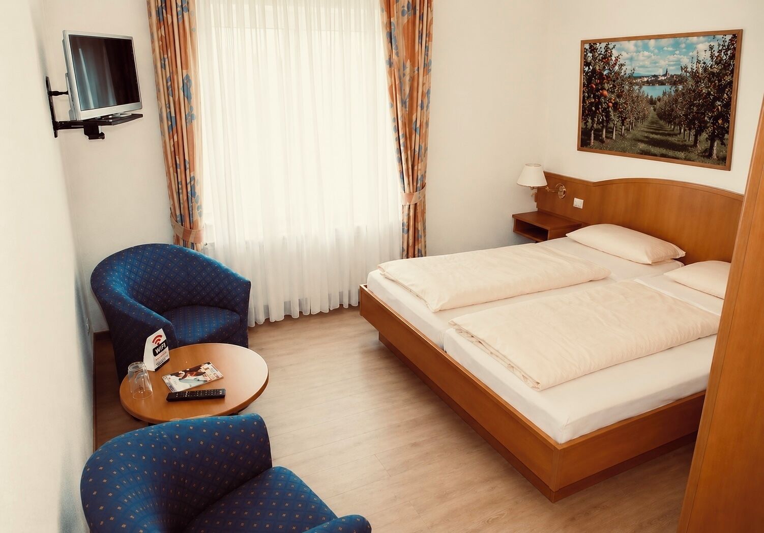 A hotel room with a bed, a TV mounted on the wall, a desk with a lamp, and a painting on the wall.