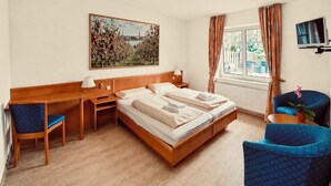 Habitación de hotel con cama, escritorio, sillas, un cuadro, televisor y ventana con cortinas.