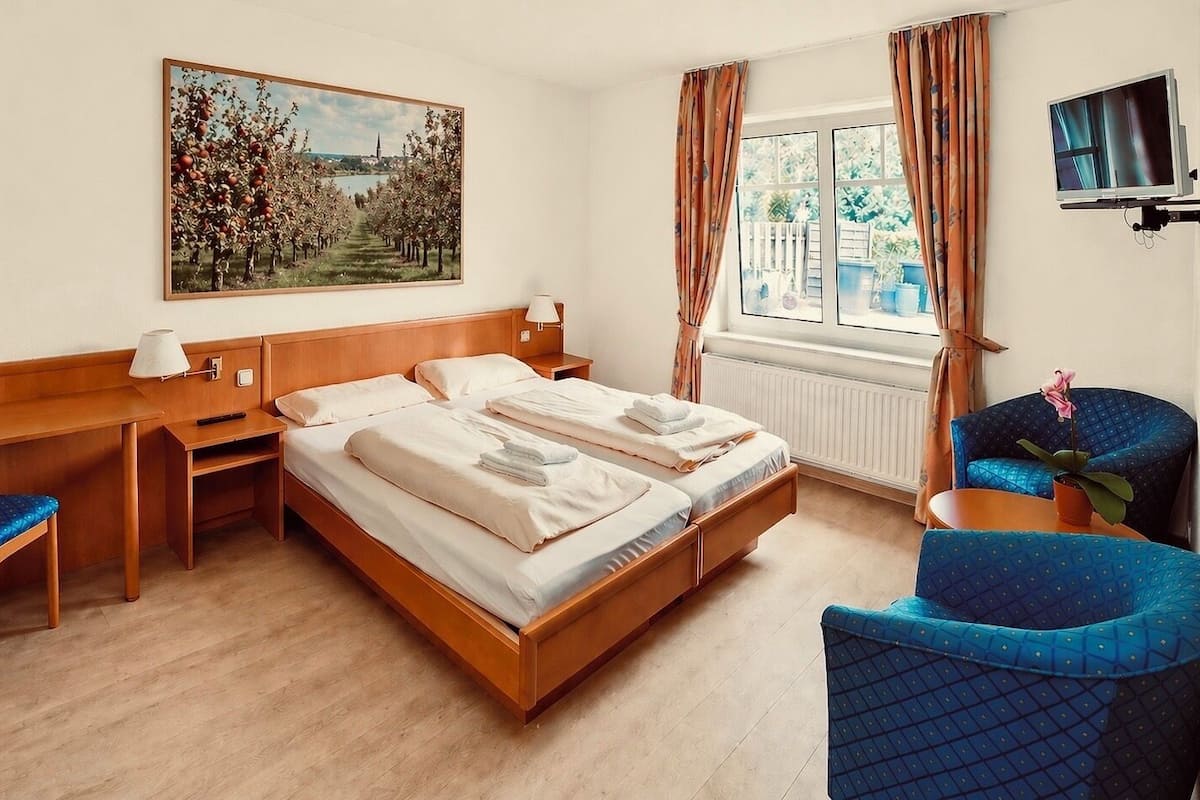 Habitación de hotel con cama, escritorio, sillas, un cuadro, televisor y ventana con cortinas.