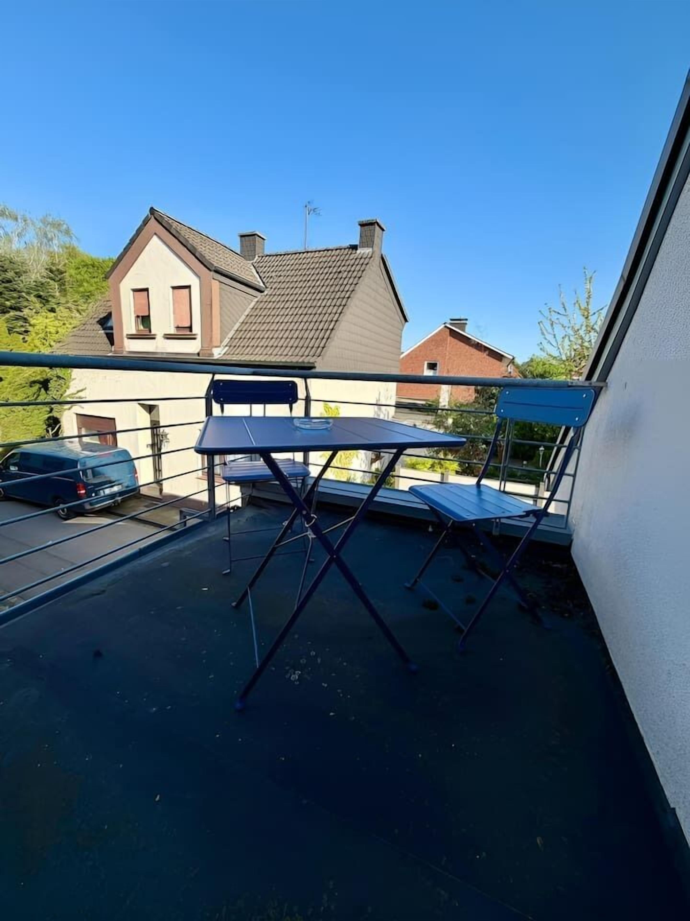 Een balkon met een tafel en stoelen, een geparkeerde auto op de achtergrond en een huis met een schuin dak.