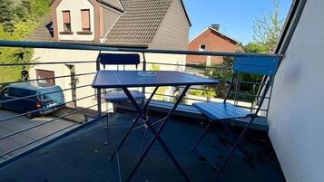 Een balkon met een tafel en stoelen, een geparkeerde auto op de achtergrond en een huis met een schuin dak.