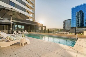A rooftop pool with lounge chairs, a small table, and a glass building in the background. - Elevated 3BR Stay - Views, Amenities, Parking (Newcastle West)