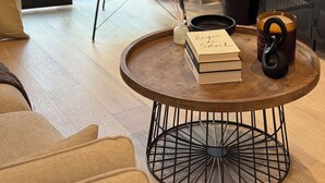 Un salon moderne avec un fauteuil en cuir, une table basse ronde ornée de livres et d’une bougie, une plante en pot et une vue à travers une porte vitrée.
