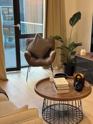 Ein modernes Wohnzimmer mit einem Ledersessel, einem runden Couchtisch mit Büchern und einer Kerze, einer Topfpflanze und Blick durch eine Glastür.