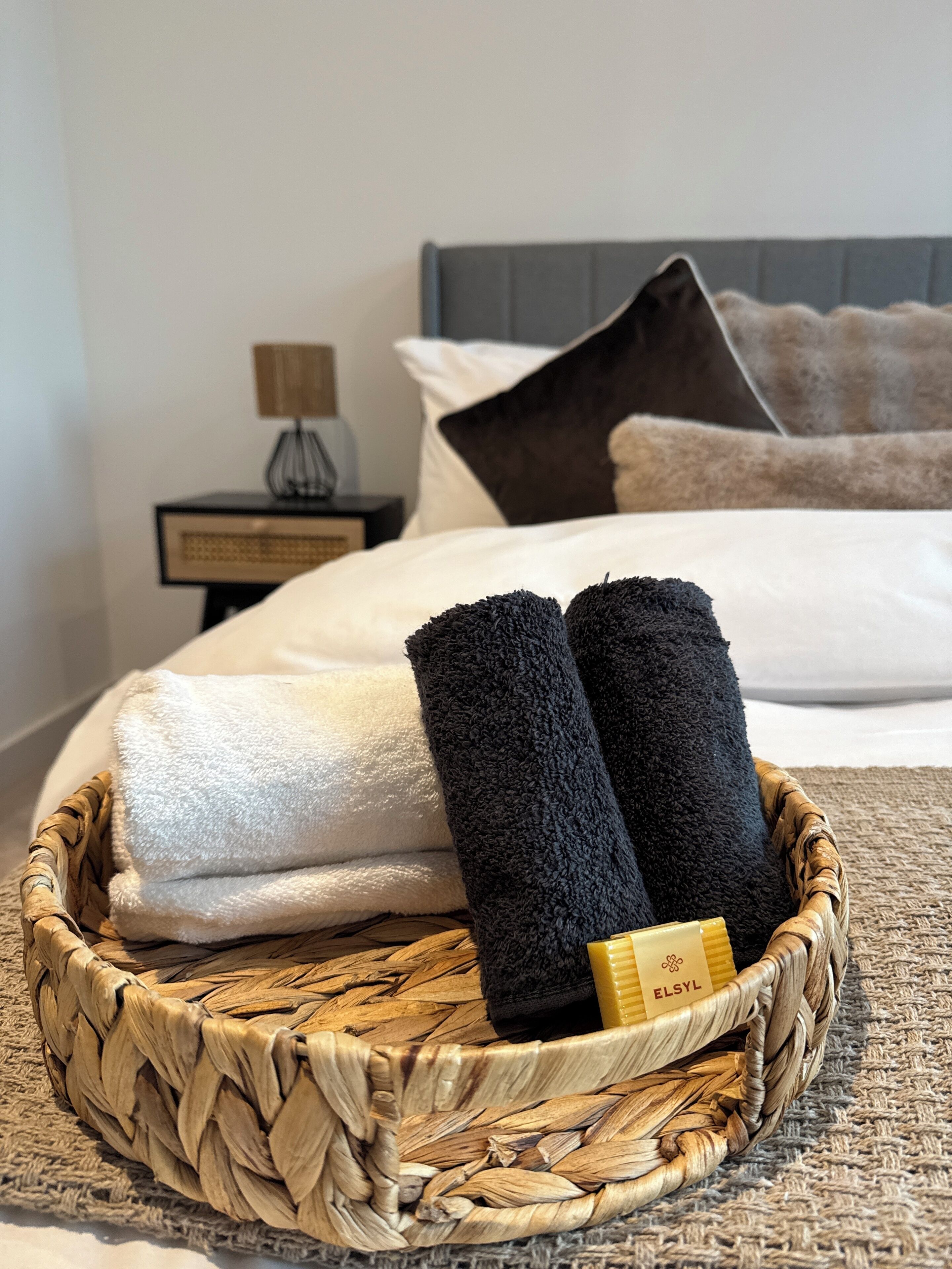 A neatly made bed with a white and black towel, a small bedside table with a lamp, and a woven basket with toiletries.