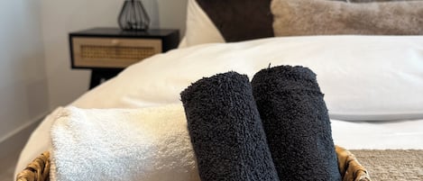 A neatly made bed with a white and black towel, a small bedside table with a lamp, and a woven basket with toiletries.
