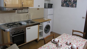 Une cuisine compacte équipée d’une cuisinière, d’un évier, d’un four à micro-ondes et d’un lave-linge. On y trouve une table à manger recouverte d’une nappe à motifs floraux, ainsi qu’une chaise.
