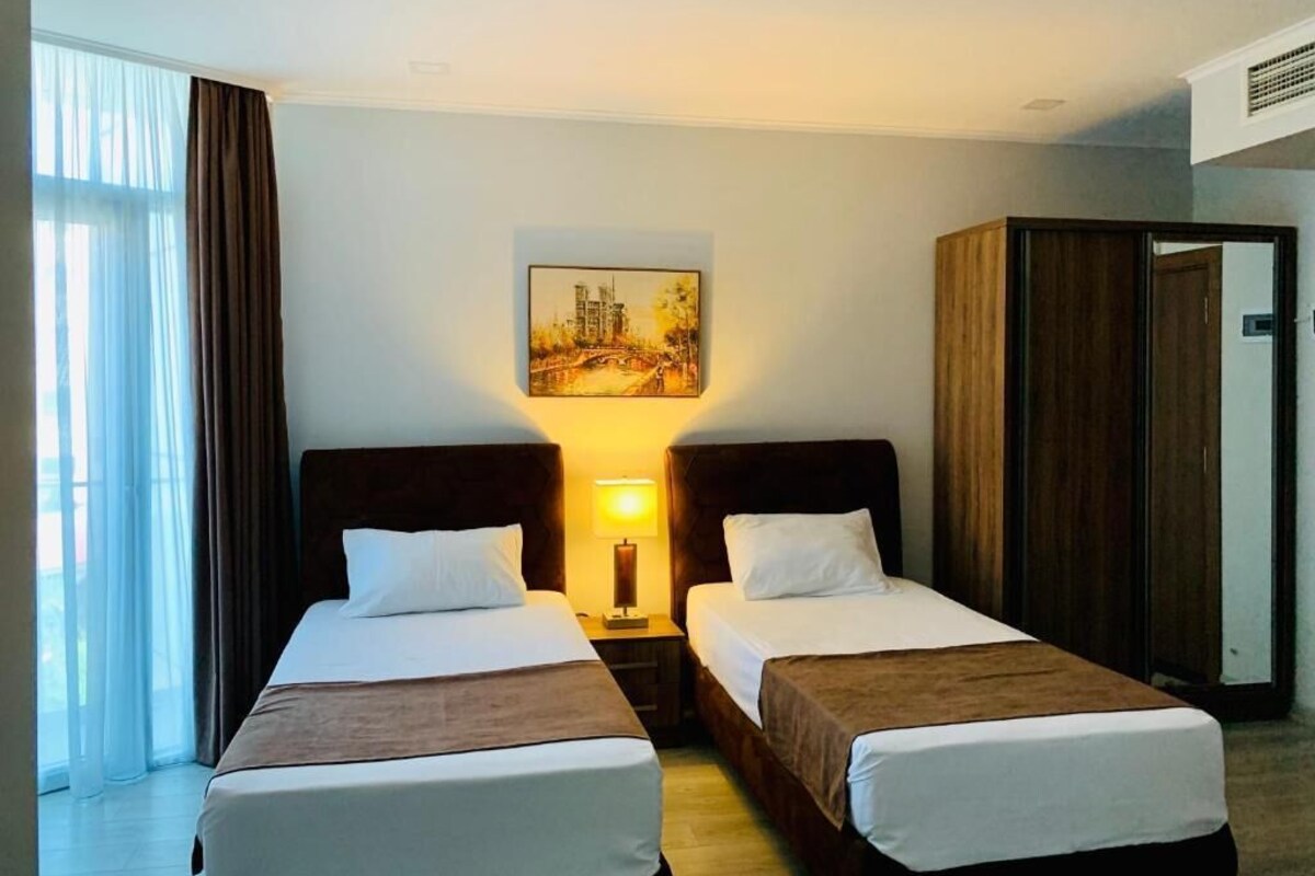 A hotel room with two beds, a nightstand with a lamp, a painting on the wall, and a wardrobe.