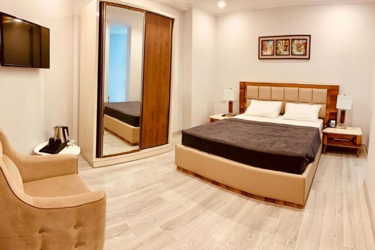 A hotel room with a bed, a chair, a TV, a bedside table, and a large mirror.