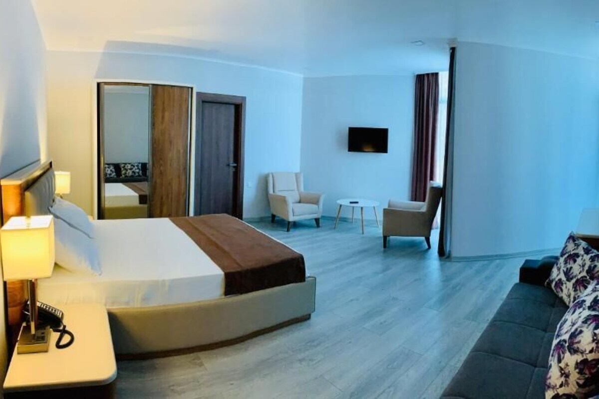 A hotel room with a bed, a sofa, a chair, a small table, a TV, and a mirror.