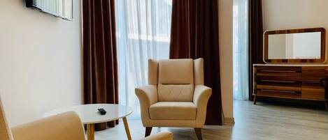 Quarto de hotel com sofá, poltrona, mesa pequena, televisão, espelho e cómoda de madeira.