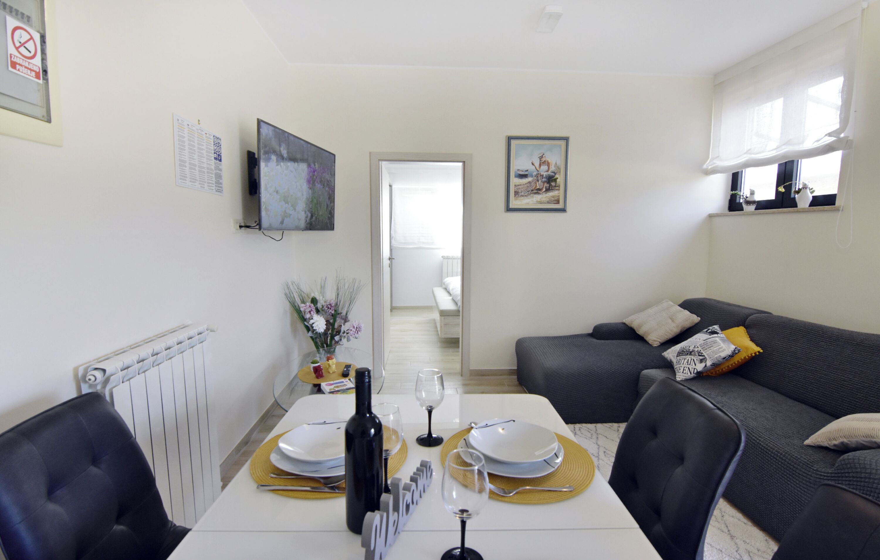 Zona pranzo con tavolo apparecchiato per due, una poltrona, una televisione e una porta che dà su un'altra stanza.