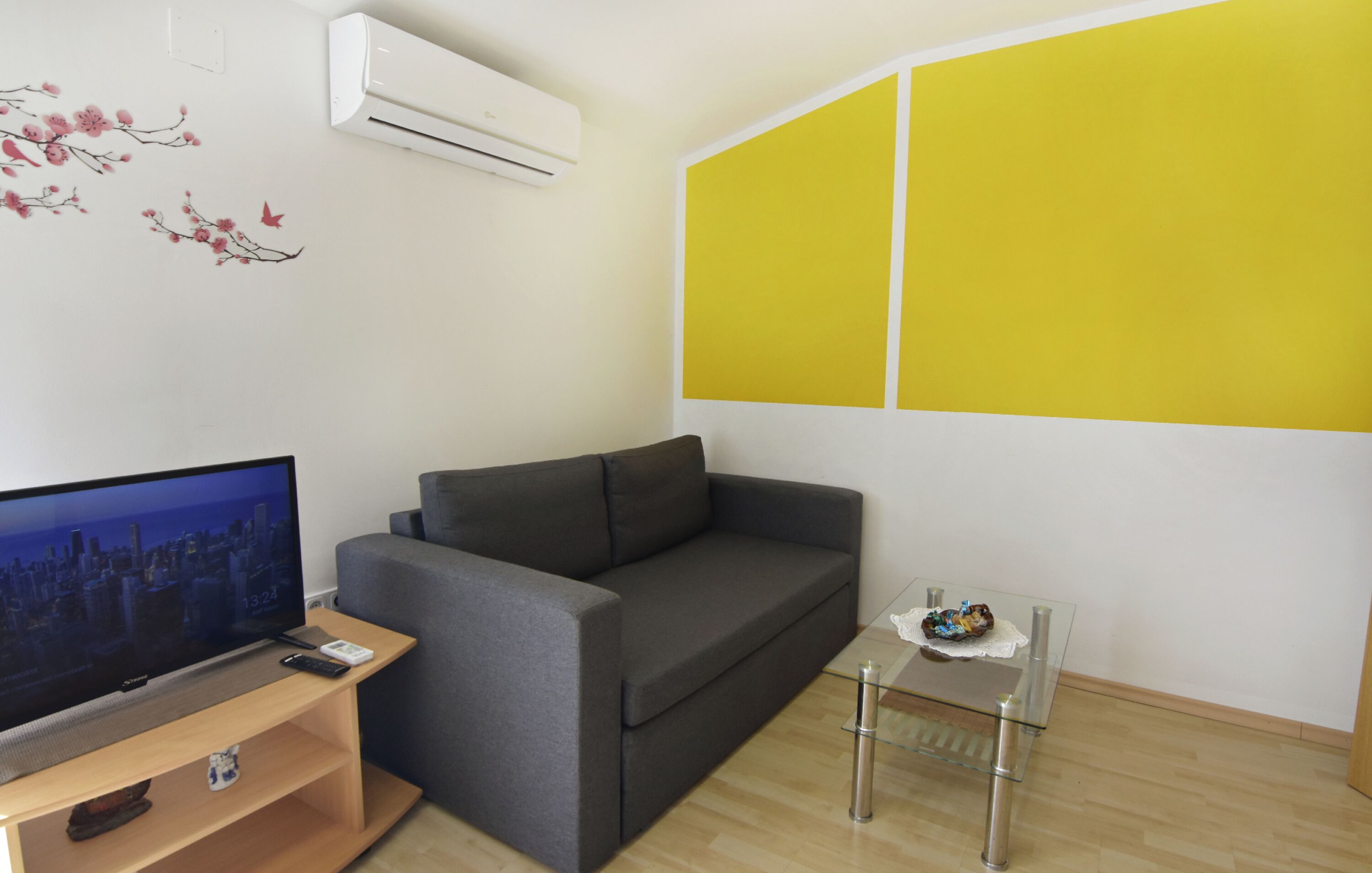 Ein Wohnzimmer mit Flachbildfernseher, grauem Sofa, Glastisch und einer gelben Akzentwand.