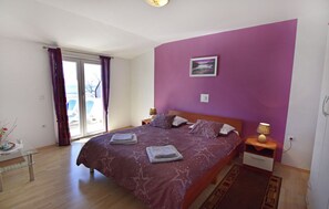 A bedroom with a large bed, a nightstand with a lamp, a glass table with a vase, and a view of the outdoors through a sliding door. - Comfortable vacation flat in a great area (Zambratija)