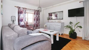 Ein Wohnzimmer mit einer Couch, einem Tisch, einem Sessel, einem Fernseher, einem Spiegel und einer Pflanze.