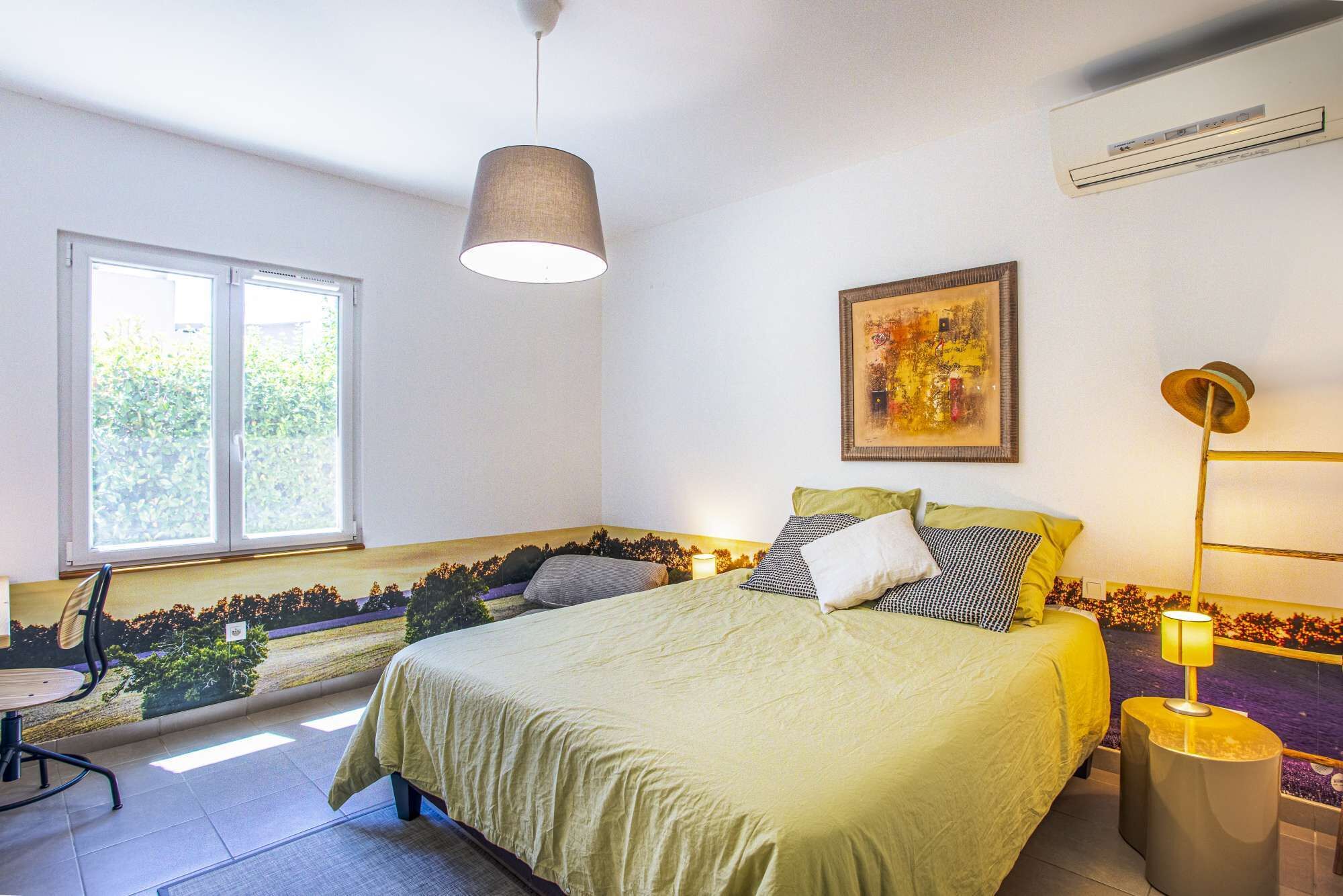 A bedroom with a bed, a window, a painting, an air conditioner, and a lamp.