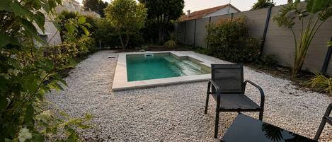 Una piscina rectangular con un área de grava alrededor, una silla negra y una mesa.