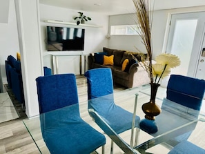 Uma sala de estar moderna com uma mesa de jantar de vidro, cadeiras azuis, um sofá com almofadas, uma televisão de tela plana e uma planta.