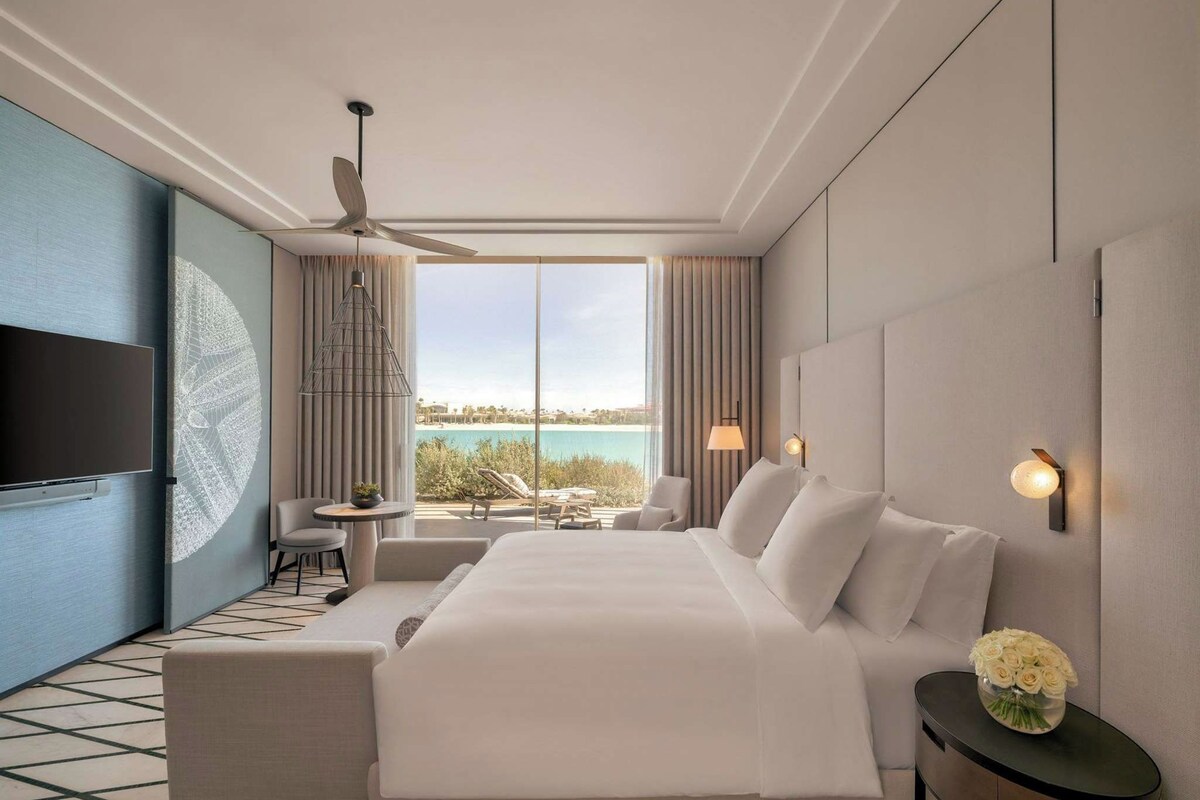 A hotel room with a large bed, a television, a ceiling fan, a view of the sea, and a small table with a chair.