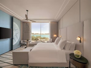 A hotel room with a large bed, a television, a ceiling fan, a view of the sea, and a small table with a chair.