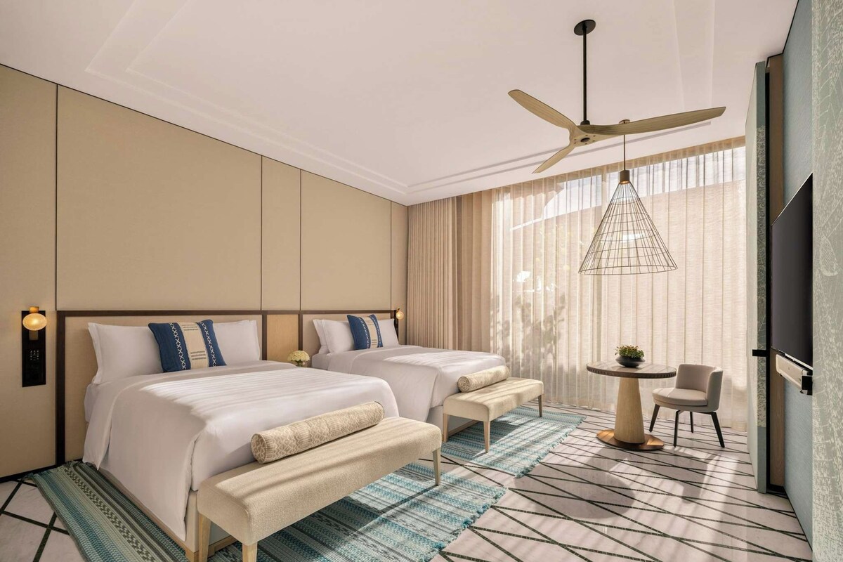 A hotel room with two beds, a ceiling fan, a small table with a chair, and a TV.