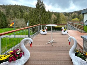 A deck with a table and two swan sculptures, surrounded by a railing and a forested backdrop. - 2 Person Holiday Park Home in Erlbach (Markneukirchen)