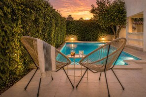 Area piscina con due sedie in vimini e un tavolo, circondata da un siepe e un edificio con un balcone.