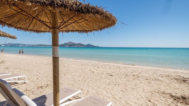 A beach with a thatched umbrella, white lounge chairs, and a clear view of the sea and distant mountains. - Spacious 5-bedroom en-suite villa with pool in stunning Playa de Muro with AC (Muro)