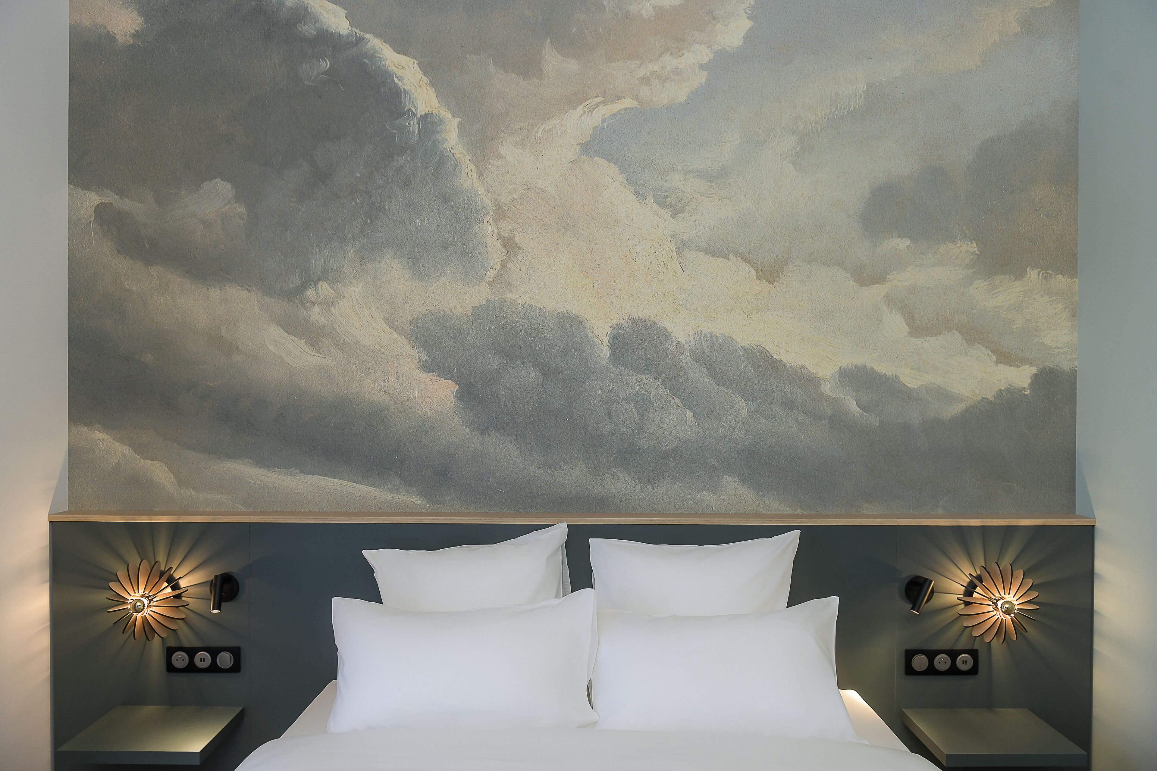 Una cama con ropa de cama blanca, dos mesitas de noche con lámparas de pared y un cuadro grande con motivo de nubes.