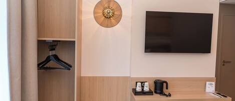 A hotel room with a wooden wardrobe, a wall-mounted TV, a desk with a coffee maker, and a hanging coat rack.
