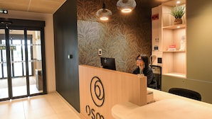 A reception desk with a logo, a plant on a shelf, and a person seated behind the desk. - The Originals Boutique Osmo Hôtel (Chantepie)