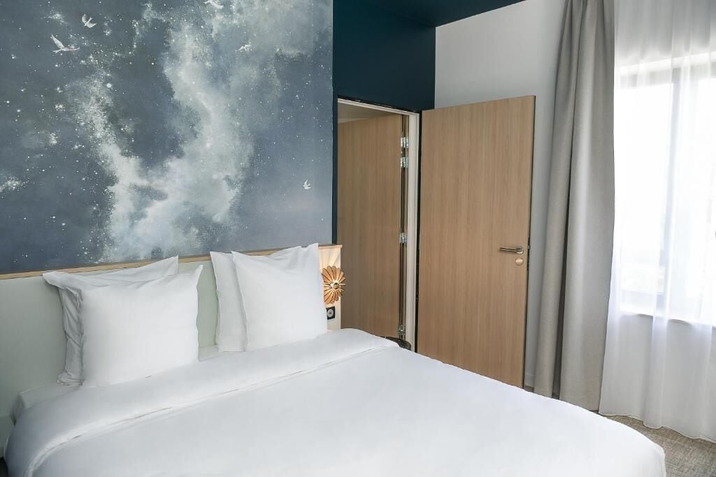 Une chambre d’hôtel avec un lit, une table de chevet, un mur orné d’une fresque sur le thème de la galaxie, une porte en bois et une fenêtre avec des rideaux.