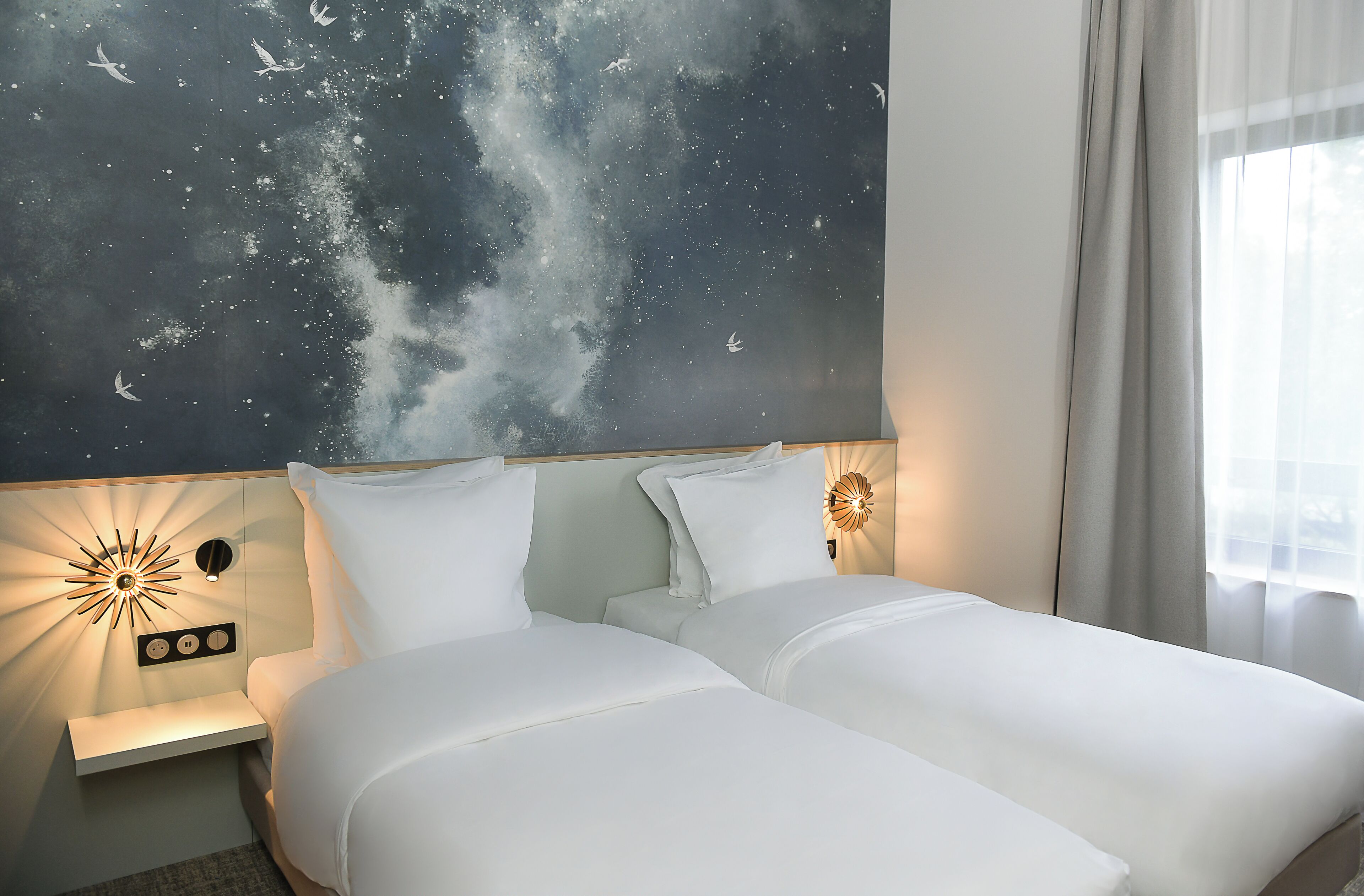 Habitación de hotel con dos camas, una mesita de noche, una lámpara de pared y un mural grande con un cielo estrellado.
