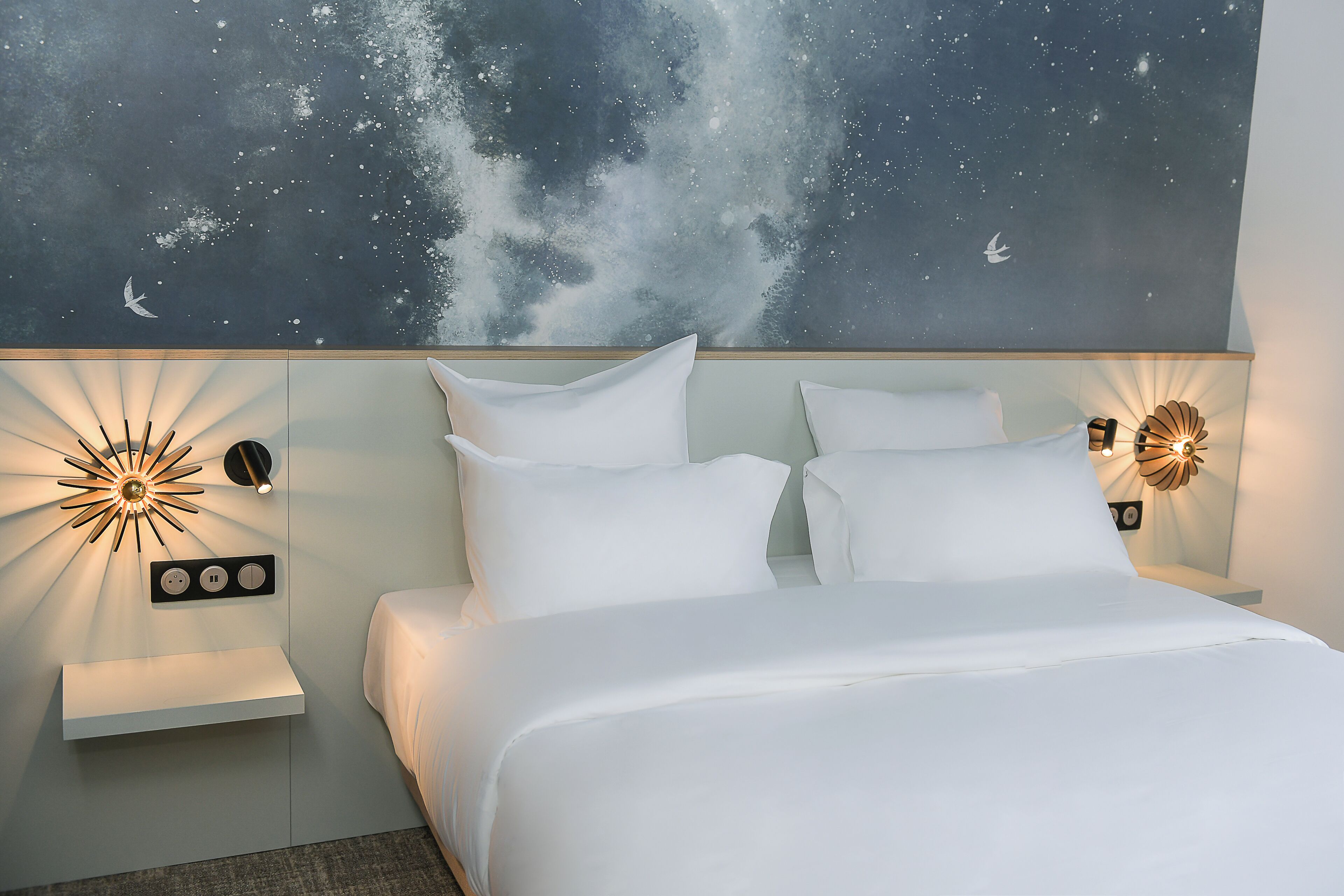 A bedroom with a bed, nightstands, wall-mounted lights, and a starry sky mural.