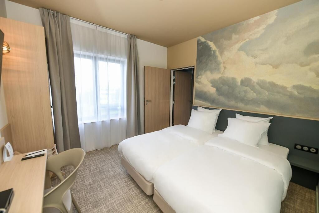 A hotel room with a large bed, a desk, and a chair. There is a painting of clouds on the wall.