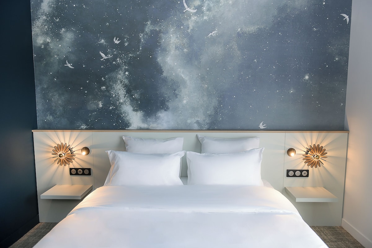 Ein Schlafzimmer mit einem Bett, zwei Nachttischen, einer Wandleuchte und einer großen Wandkunst, die einen Nachthimmel mit Vögeln zeigt.