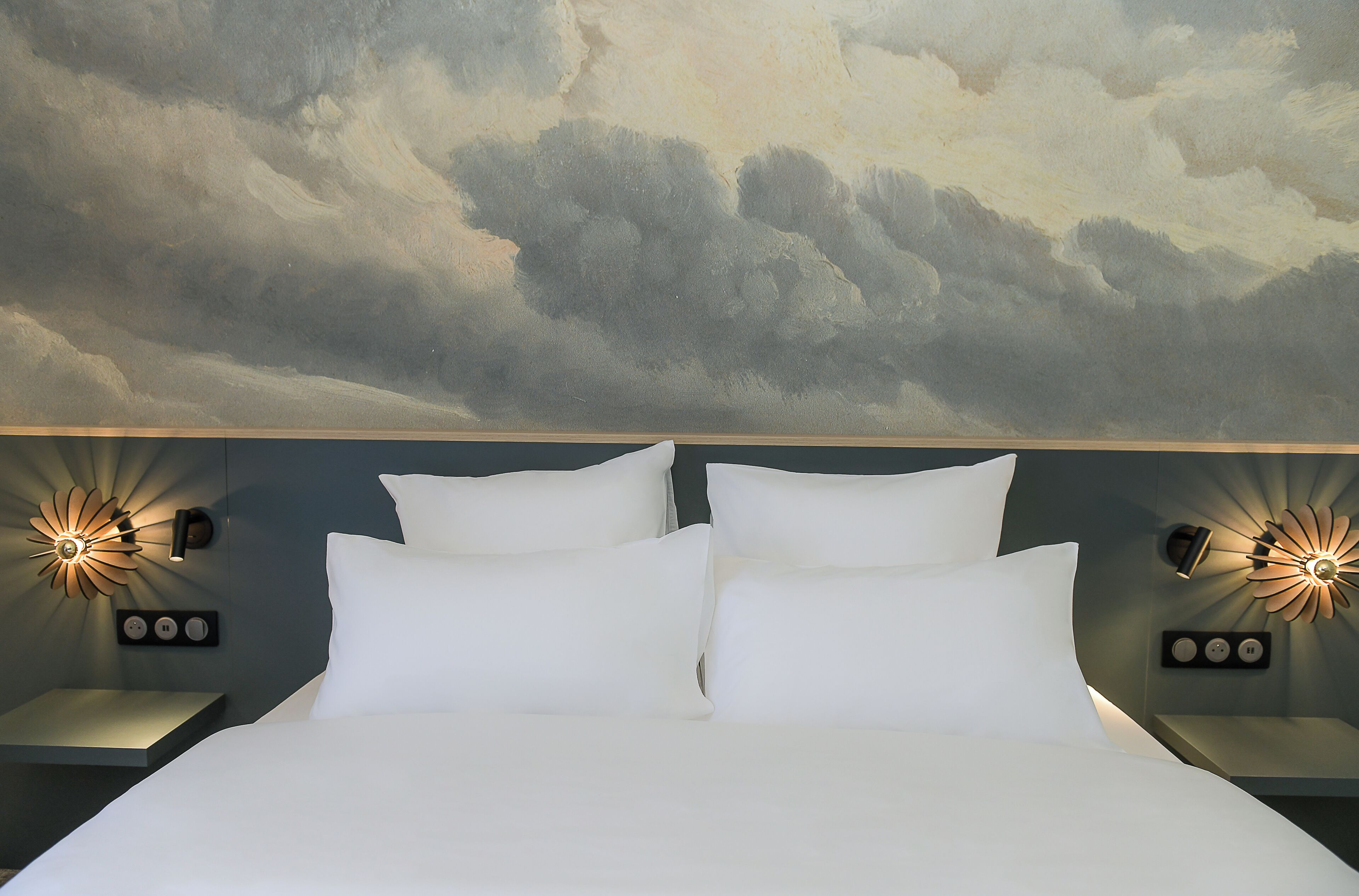Un lit avec du linge de lit blanc, deux tables de chevet avec des lampes fixées au mur, et une fresque représentant des nuages peinte au plafond.