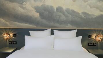 Un lit avec du linge de lit blanc, deux tables de chevet avec des lampes fixées au mur, et une fresque représentant des nuages peinte au plafond.