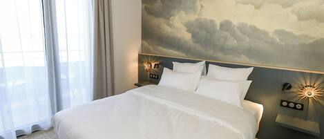 A hotel room with a large bed, a nightstand with a lamp, a wall-mounted light, and a view of the sky through the window.