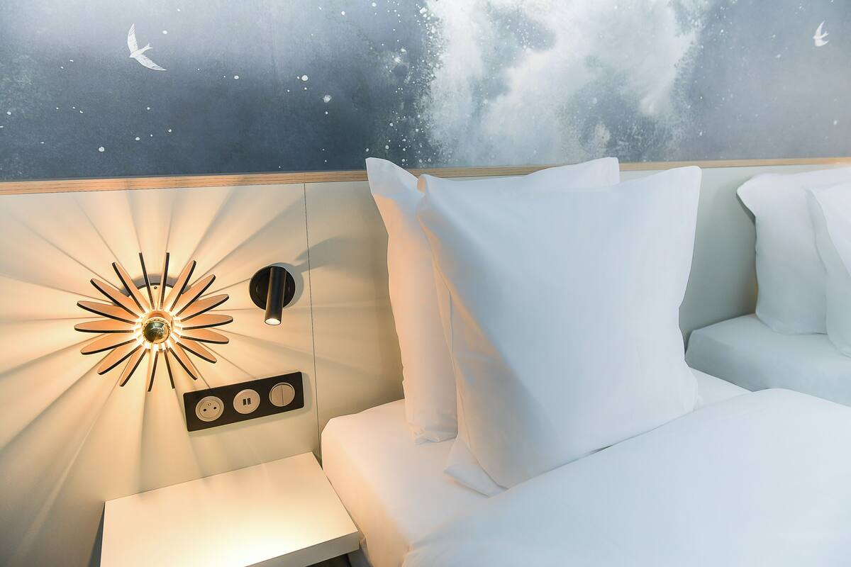 Ein Schlafzimmer mit Wandmalerei, die einen sternenübersäten Nachthimmel zeigt, einer modernen Wandleuchte und einem ordentlich bezogenen Bett mit weißer Bettwäsche.