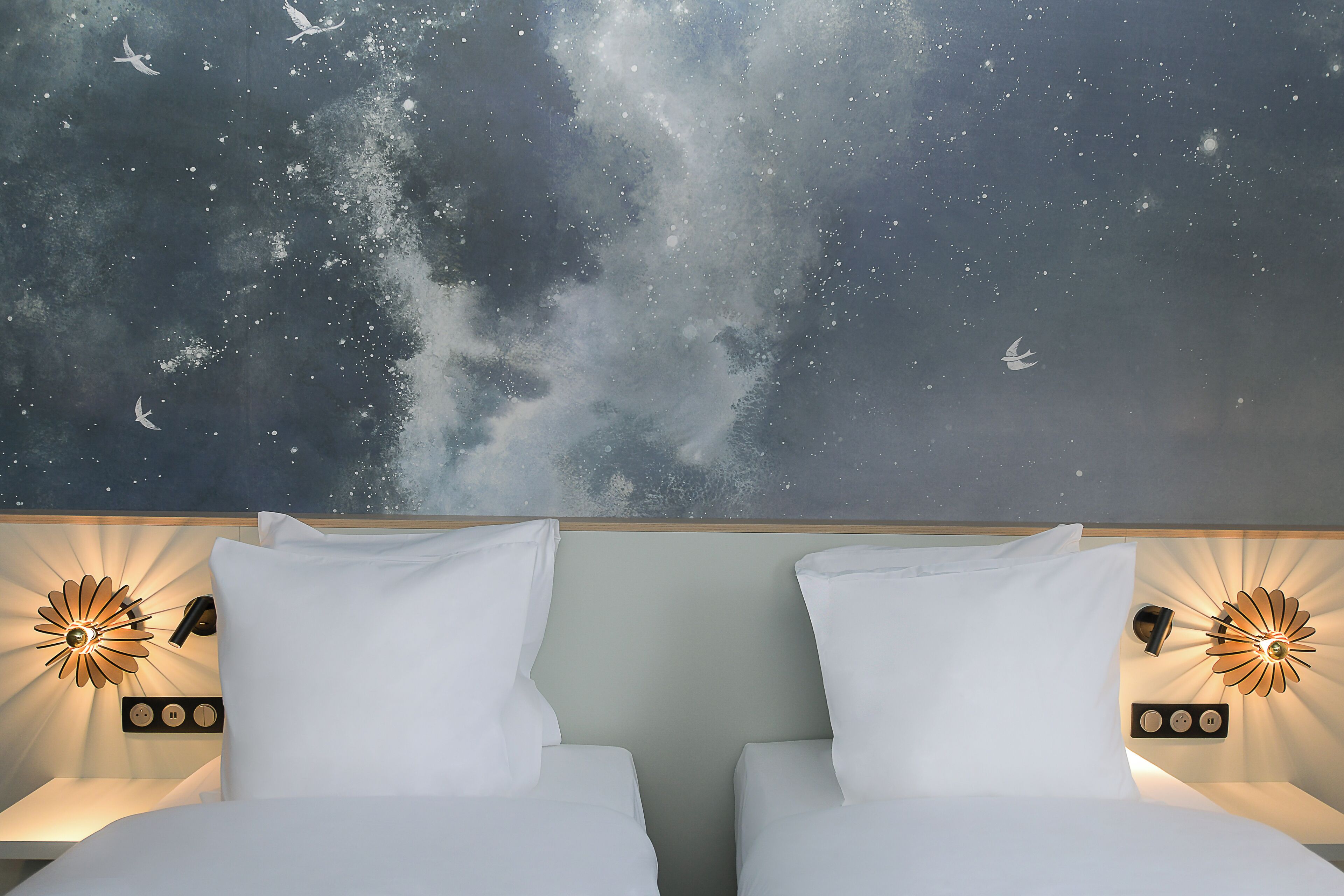 Habitación de hotel con una cama, dos mesitas de noche y un mural en la pared que representa un cielo estrellado.