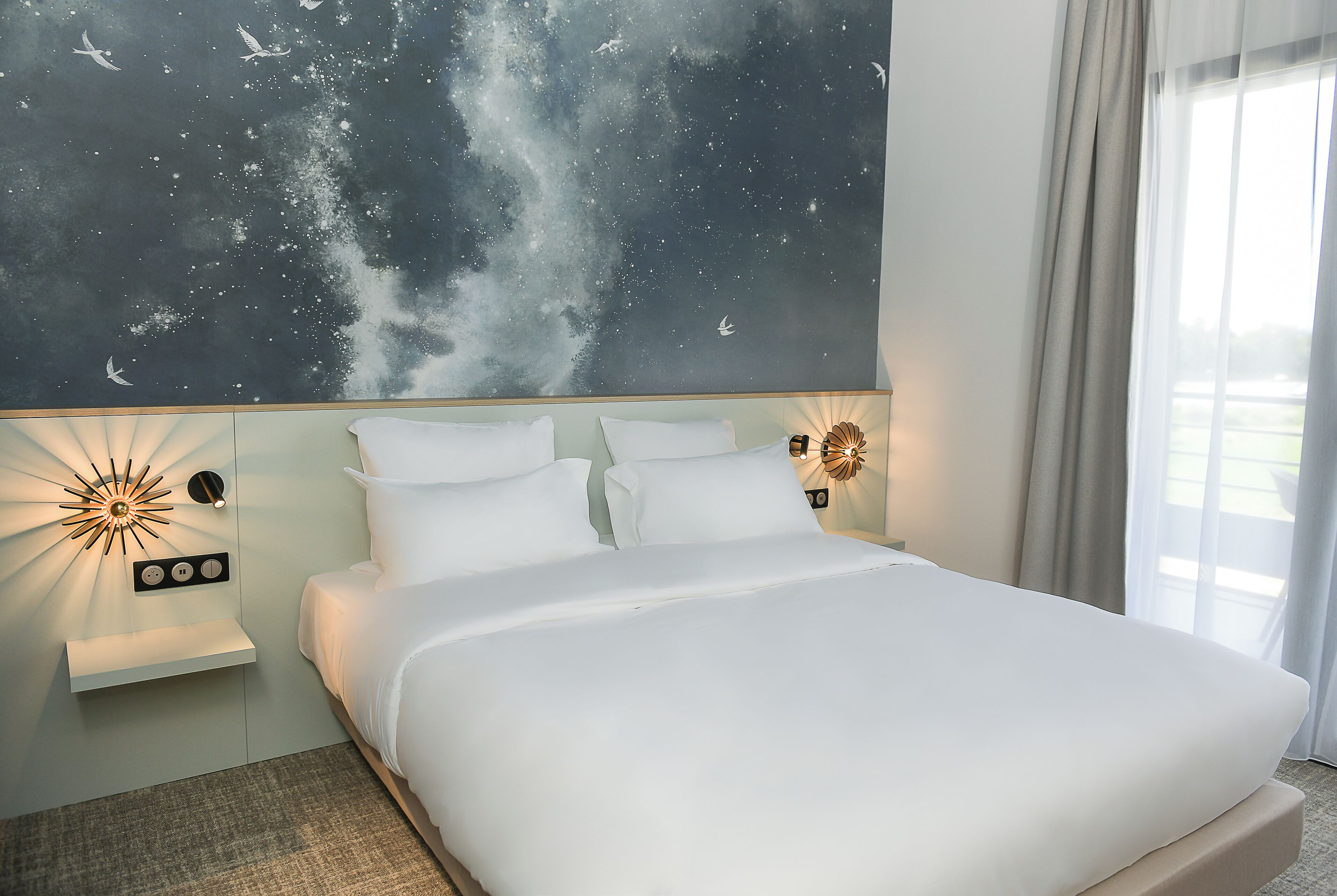 Habitación de hotel con cama, un mural del cielo nocturno, un aplique de pared y una ventana con cortinas.