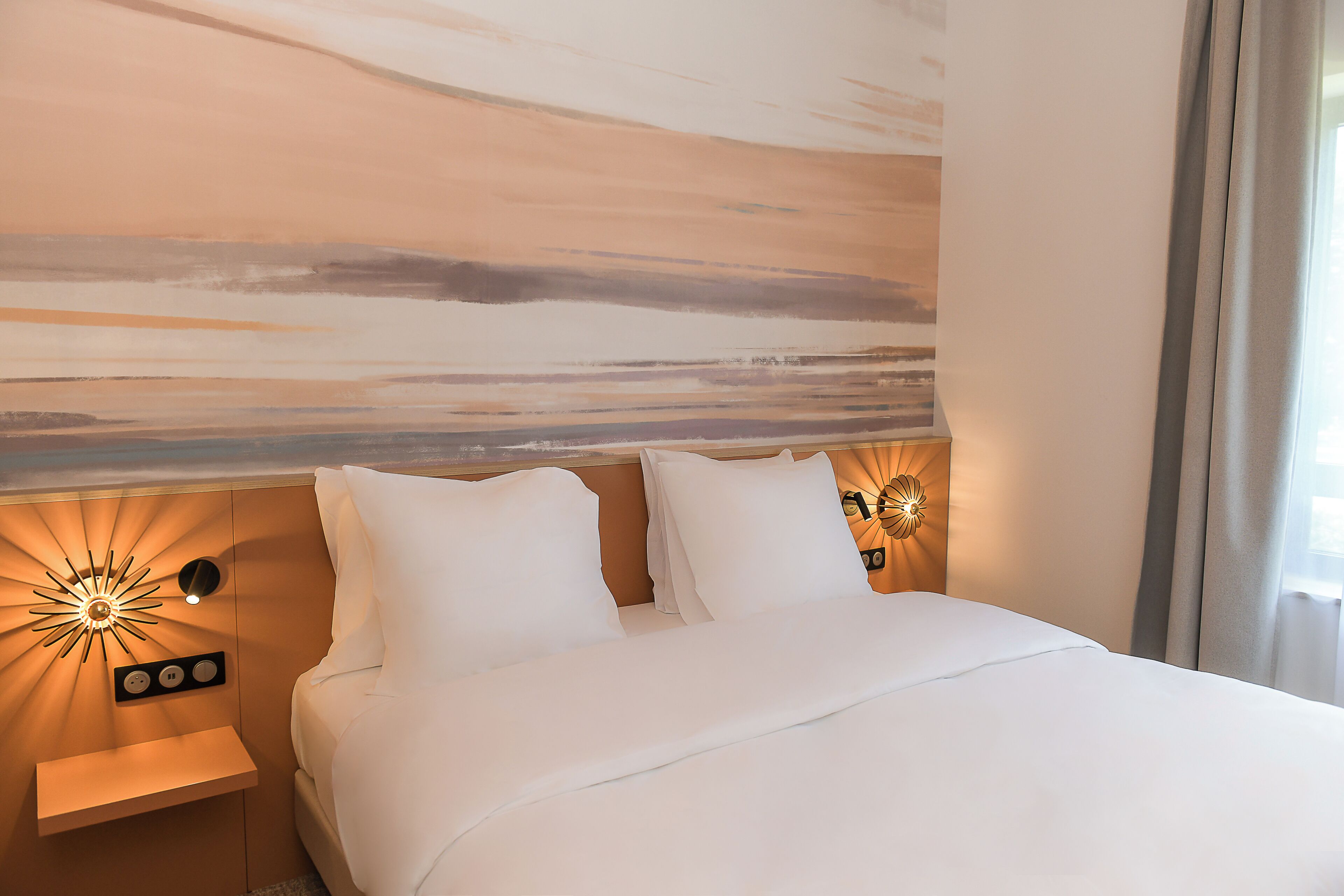 A hotel room with a bed, two bedside tables, a wall-mounted light fixture, and a view of the outdoors.