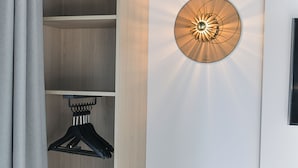 A hotel room with a wooden wardrobe, a wall-mounted light fixture, a hanging clothes rack, and a small wooden bench. - The Originals Boutique Osmo Hôtel (Chantepie)