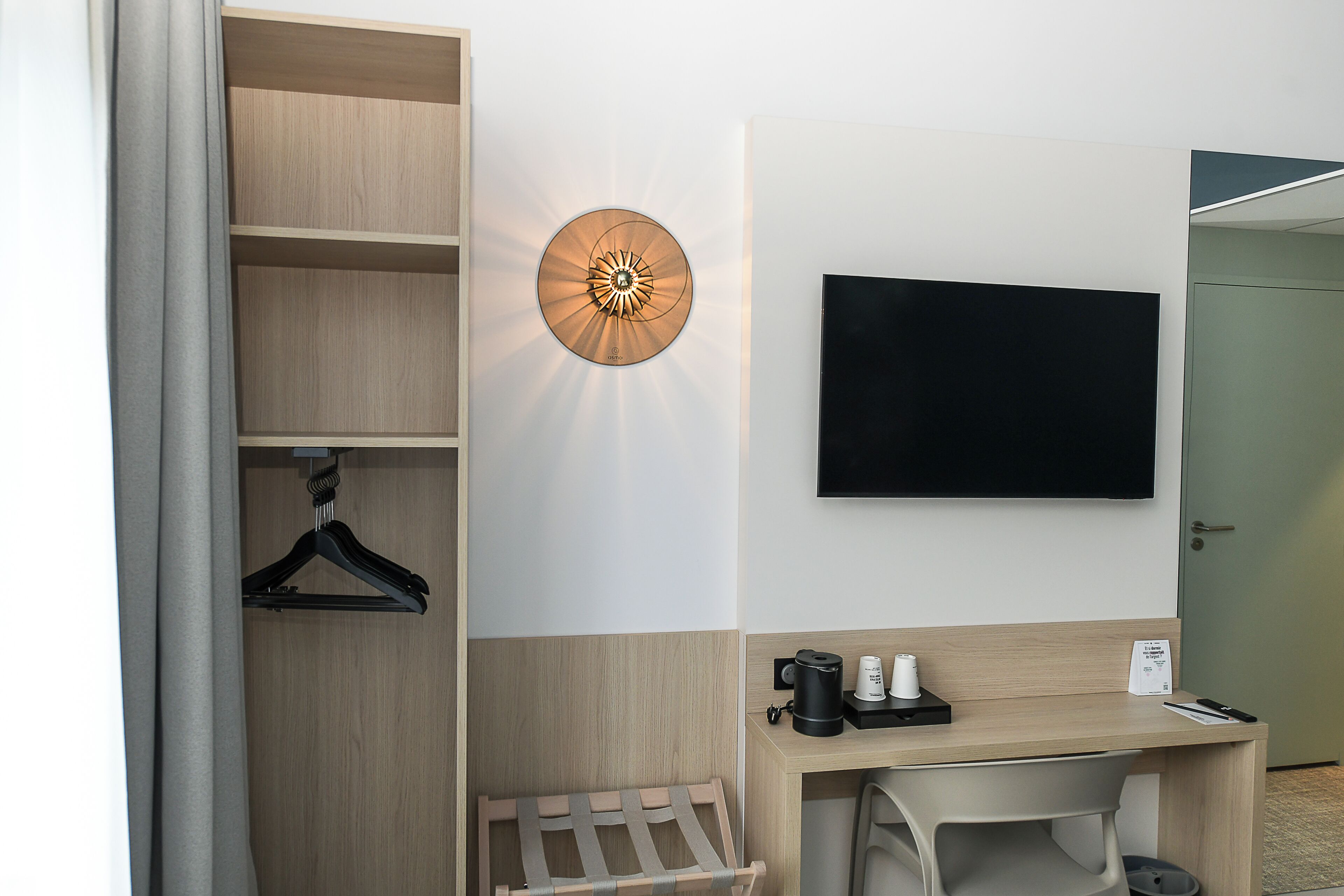 Habitación de hotel con escritorio de madera, televisor de pantalla plana, lámpara de pared, perchero y un pequeño rincón de descanso.