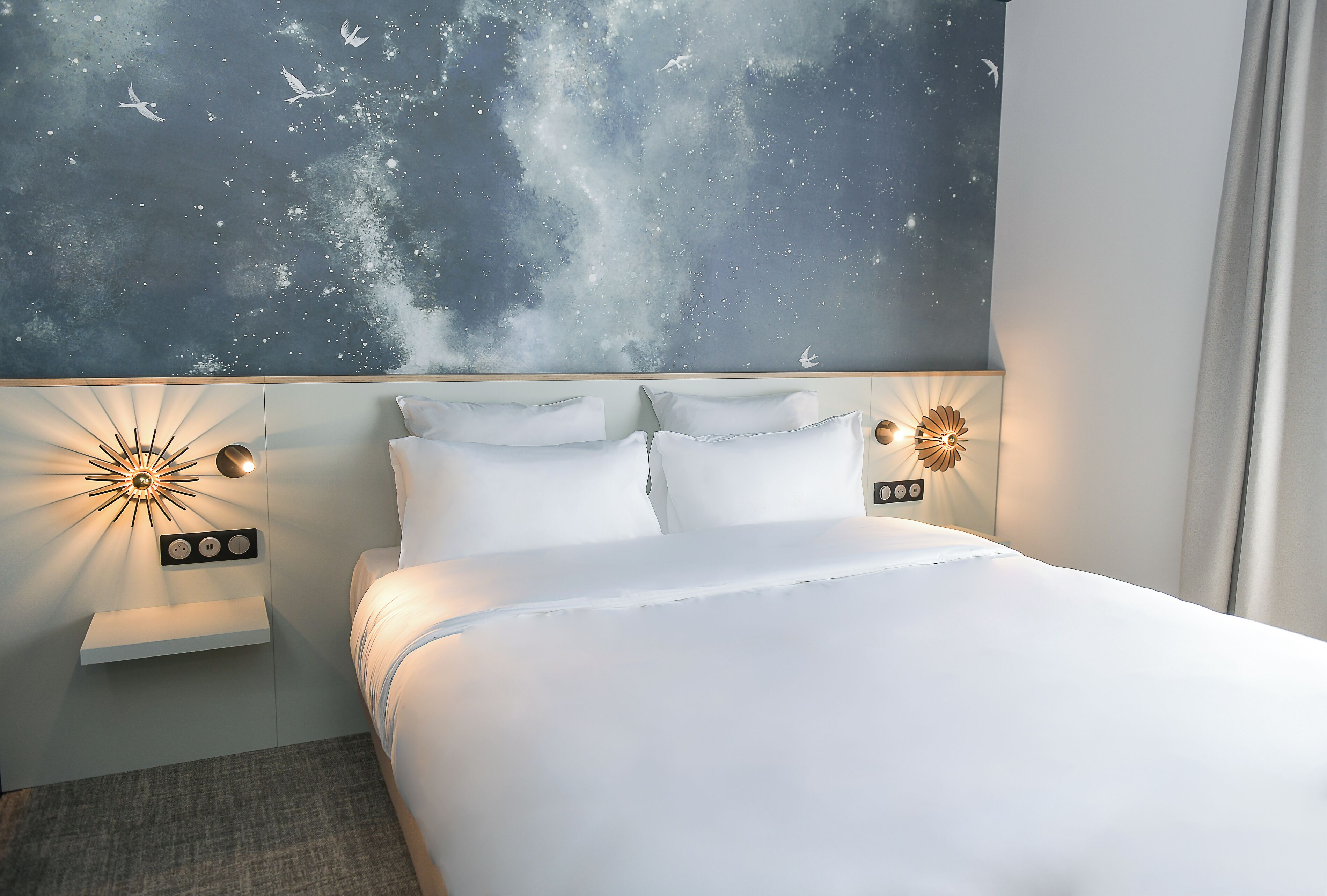 Une chambre d’hôtel moderne avec un grand lit, une fresque murale représentant un ciel étoilé, des lampes de lecture fixées au mur et une table de chevet avec une lampe.