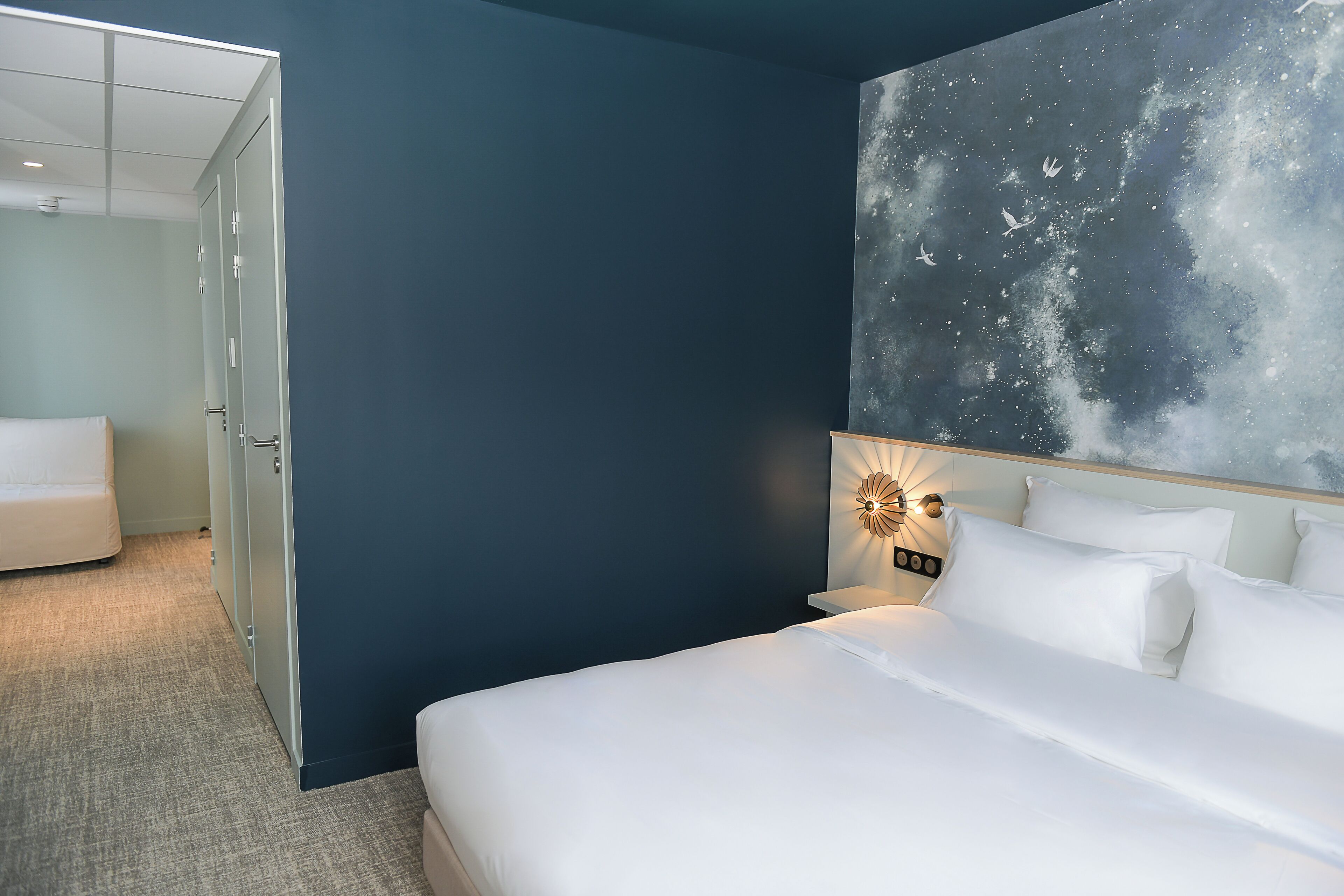 A hotel room with a bed, a nightstand, a wall-mounted light, and a door leading to another room.