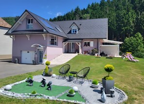 Ein zweistöckiges Haus mit Satteldach, einer Veranda und einem begrünten Vorgarten mit Stühlen und Pflanzen in Töpfen.