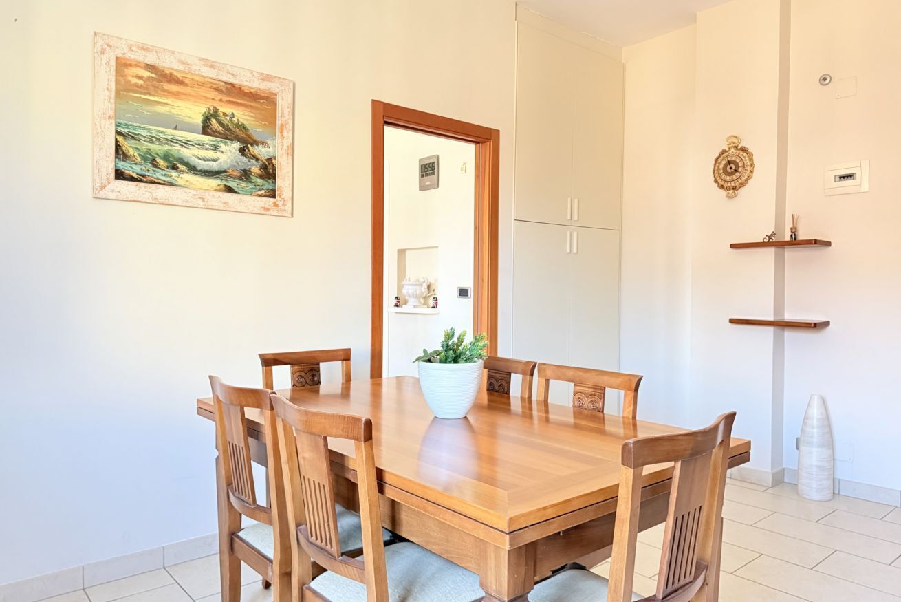 A dining area with a wooden table and chairs, a potted plant, a framed painting, and a door leading to a kitchen.