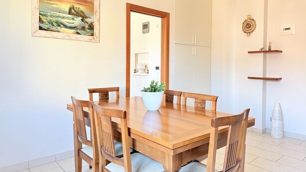 Een eetruimte met een houten tafel en stoelen, een potplant, een lijst met een schilderij en een deur naar een keuken.